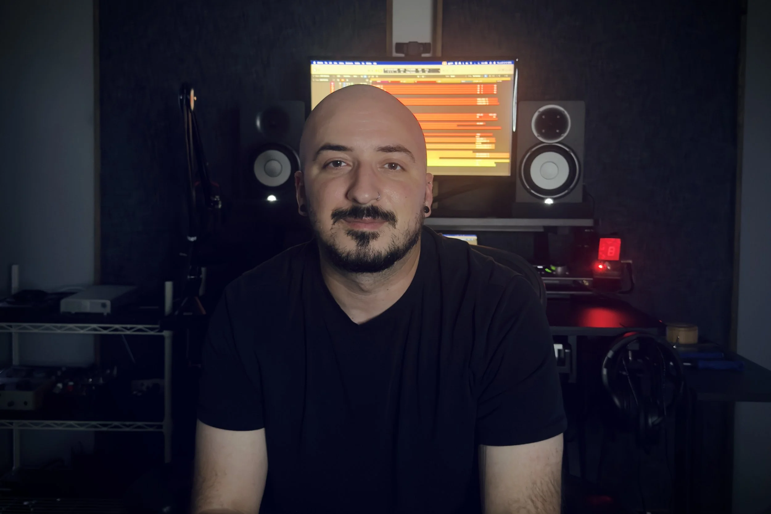 A man with a shaved head, beard, and nose piercing sitting in a home recording studio with studio monitors and a computer screen displaying audio editing software in the background.