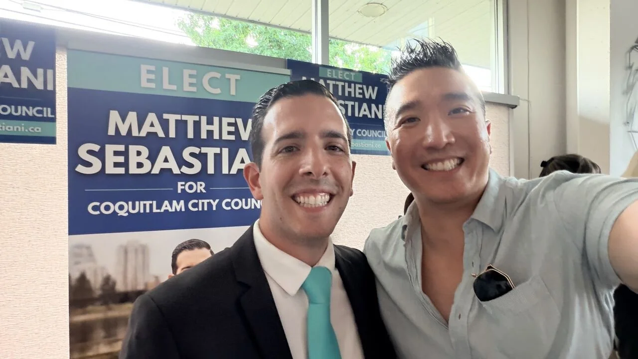 Two men smiling in front of a campaign poster that reads "Elect Matthew Sebastian for Coquitlam City Council". They are taking a selfie, with one wearing a suit and tie and the other in a casual shirt, indoors with a window behind them.