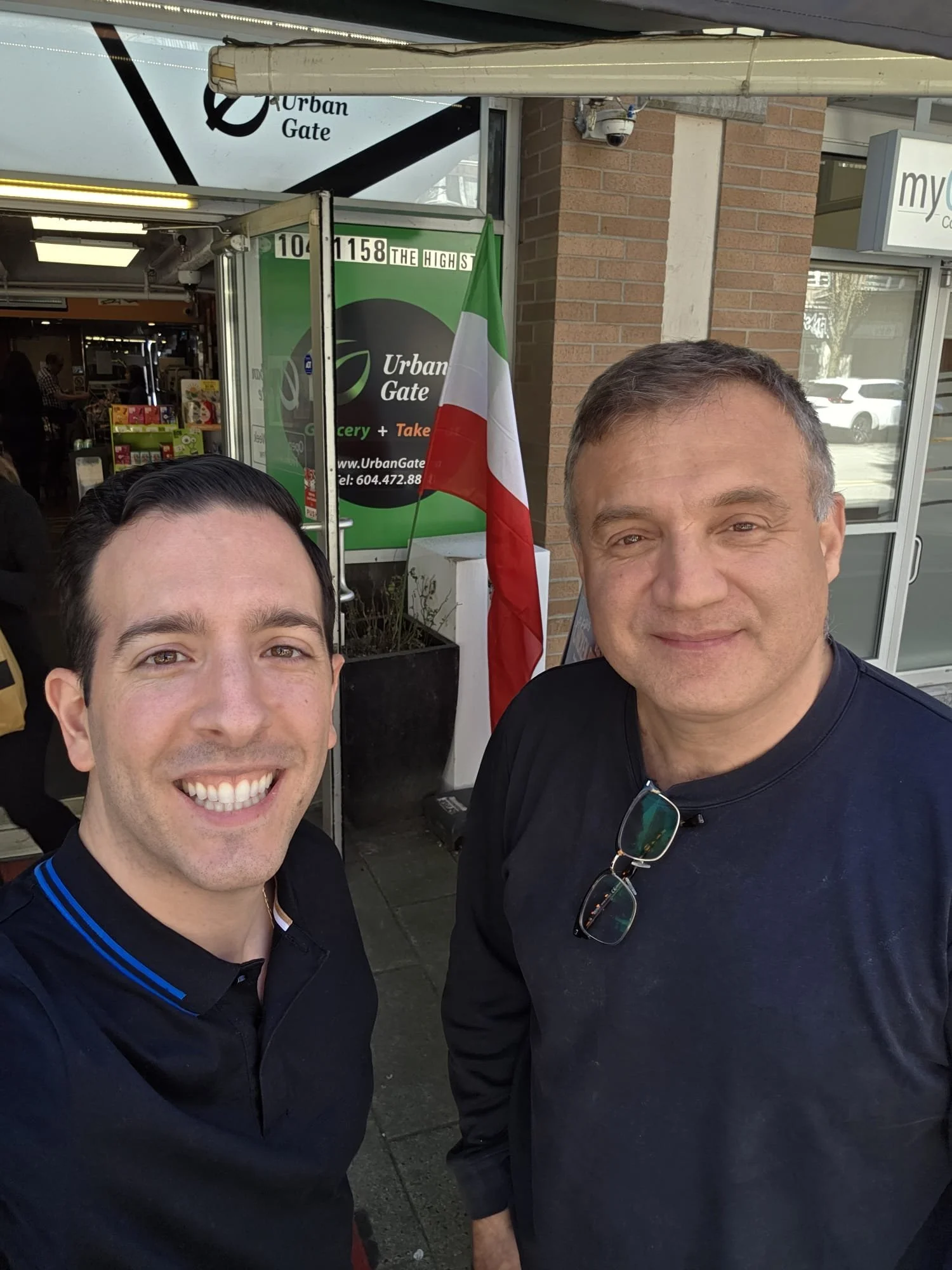 Two smiling men taking a selfie outside a store with a green Urban Gate sign and an Italian flag. The store's entrance is visible in the background.