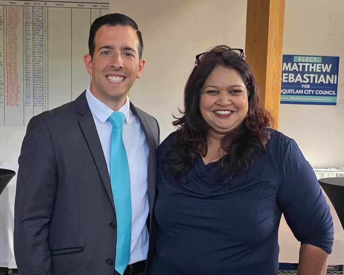 A man in a suit with a light blue tie and a woman in a dark blue top standing together, smiling for a photo indoors. There is a sign advocating for Matthew Sebastian for council in the background.