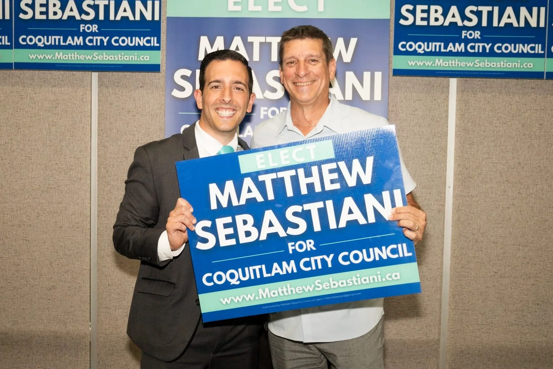 Two men smiling at an indoor campaign event, holding a blue sign that reads "Elect Matthew Sebastian for Coquitlam City Council", with campaign posters in the background.