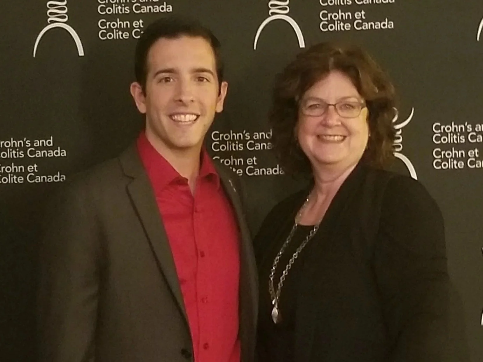 Two women standing in front of a black backdrop with white text and designs. The woman on the left is young, with short dark hair, wearing a dark blazer over a red shirt. The woman on the right is older, with curly brown hair, glasses, and wearing a black blazer and a necklace.