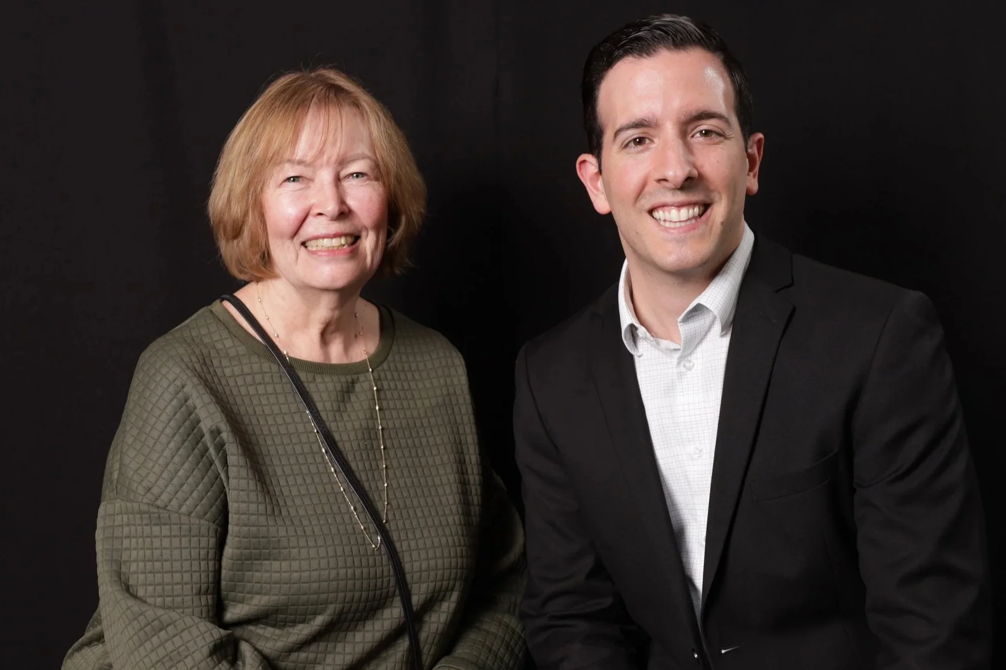 A woman with shoulder-length blonde hair, wearing a green quilted top and a long necklace, standing next to a smiling man with short dark hair, wearing a black blazer and white dress shirt, in front of a black background.