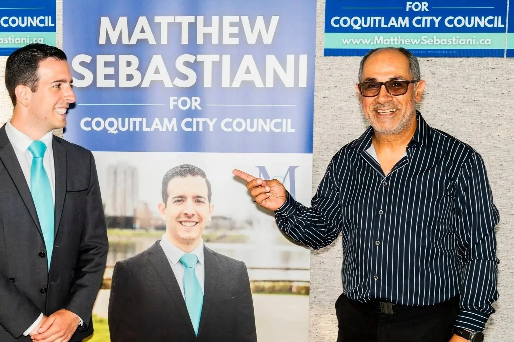 A man wearing glasses and a striped black shirt is pointing at a campaign poster for Matthew Sebastian for Coquitlam City Council, featuring images of two other men in suits with blue ties.