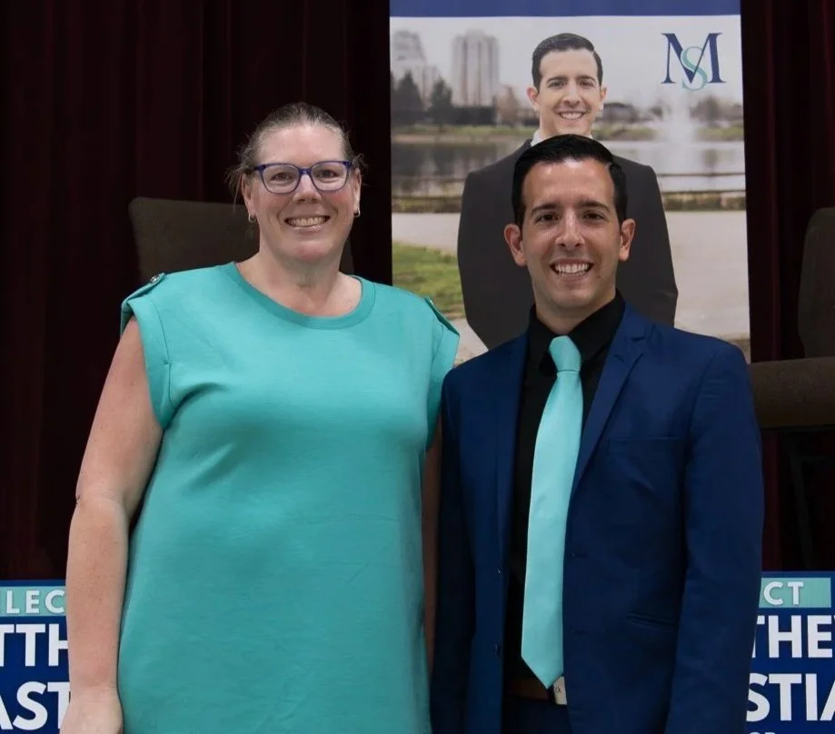 A woman and a man standing side by side, smiling, in front of a backdrop with a printed image of another smiling man and an outdoor cityscape. The woman wears glasses and a teal sleeveless top, and the man wears a blue suit with a matching tie.