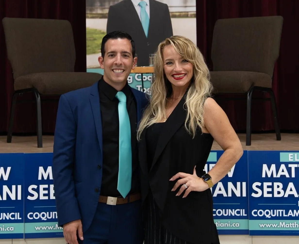 A man and woman smiling at a political event with campaign signs for Matti Sebastian in front of them. The man wears a blue suit with a black shirt and a light blue tie, and the woman wears a black sleeveless dress and has blonde wavy hair.