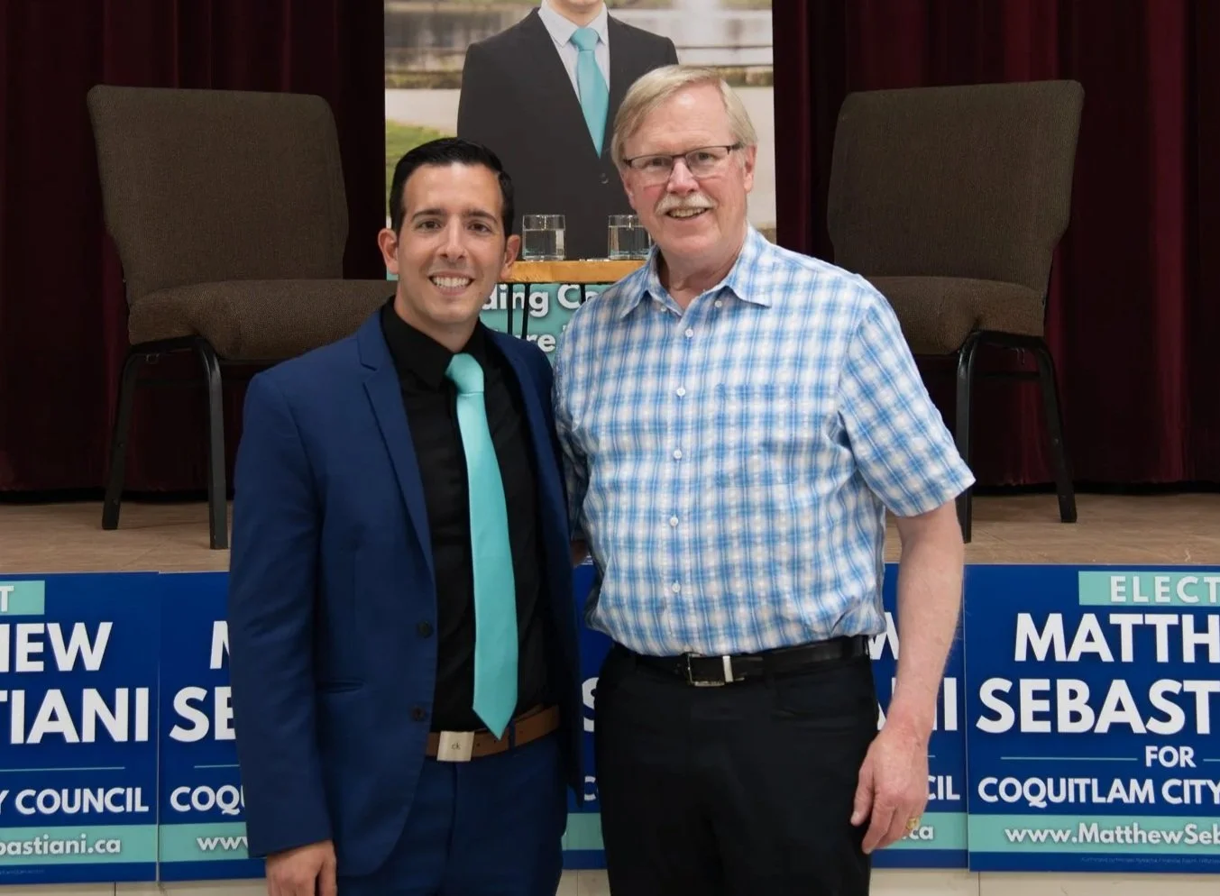 Two men standing together in front of a political campaign stage, smiling for the camera. The man on the left wears a blue suit with a matching tie, and the man on the right wears a plaid shirt. Campaign signs for Matthew Sebasti in Coquitlam City are displayed at their feet.