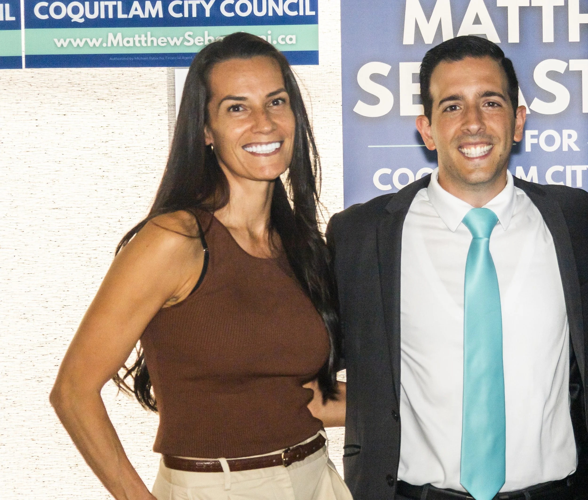 Two people standing in front of campaign signs, smiling for a photo. The woman has long dark hair, is wearing a brown sleeveless top and white pants. The man has dark hair, is dressed in a suit with a blue tie, and is smiling broadly.