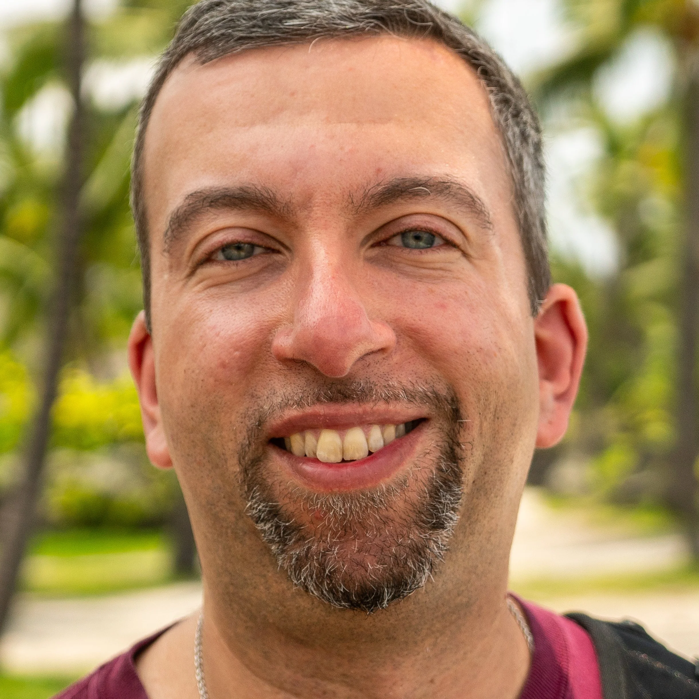 Close-up of a smiling man with short brown hair, light skin, and a beard, outdoors with blurred green trees in the background.