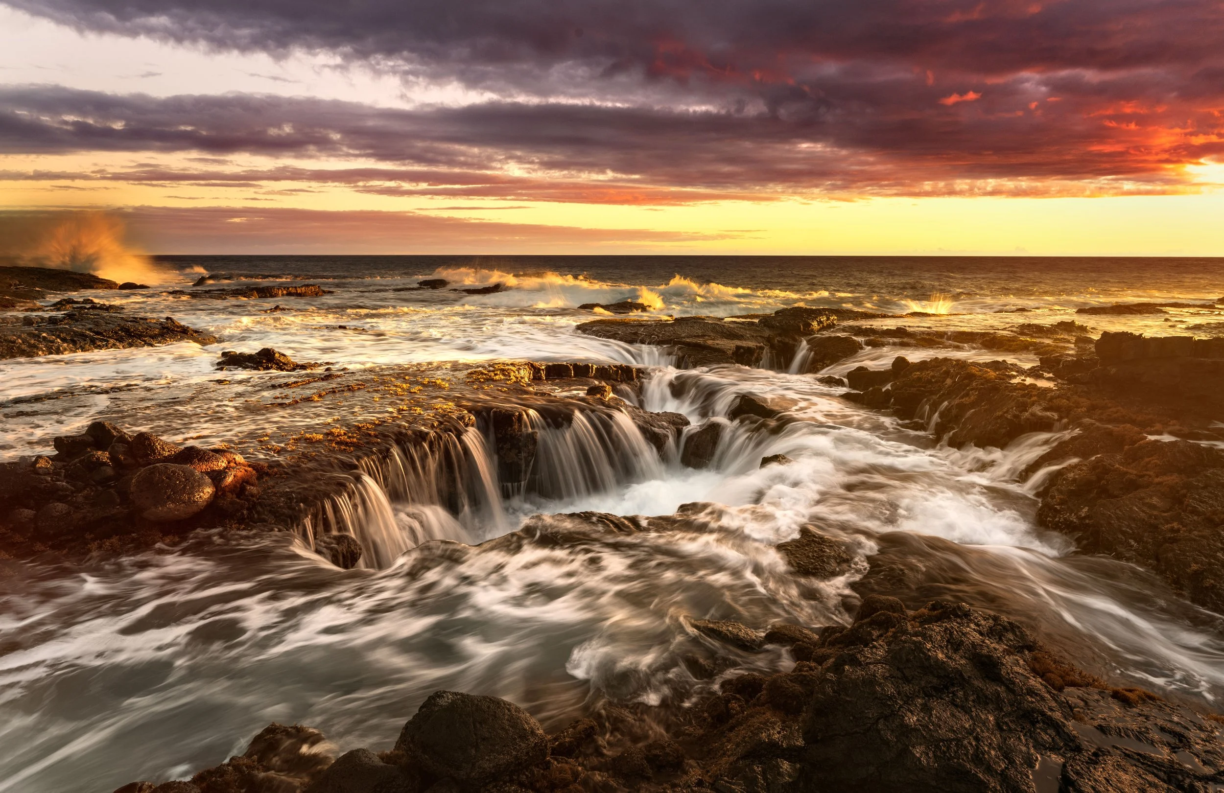 Ocean waves crashing over rocks at sunset with a colorful sky and dramatic clouds.
