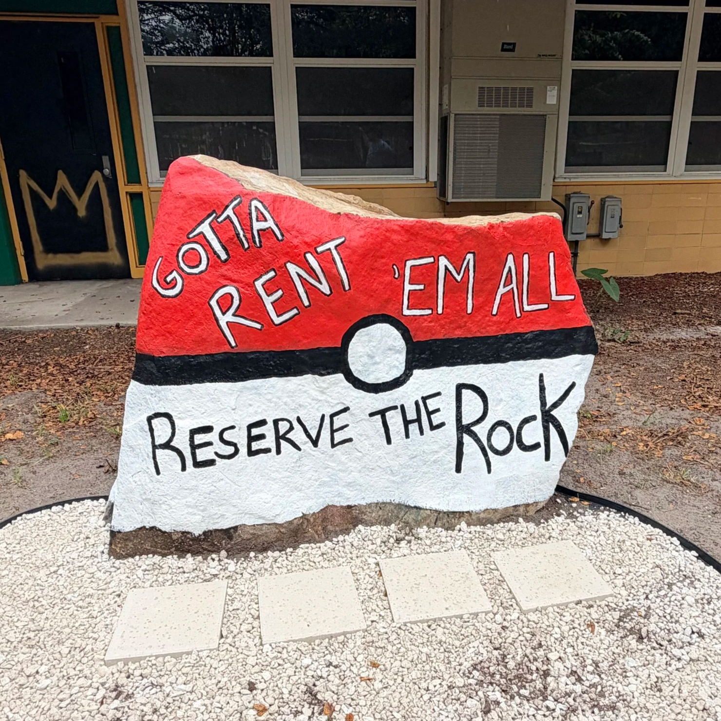 Painted rock with a red top and white bottom, styled to resemble a Poké Ball, with black outline and text that reads "GOTTA RENT 'EM ALL" and "RESERVE THE ROCK".
