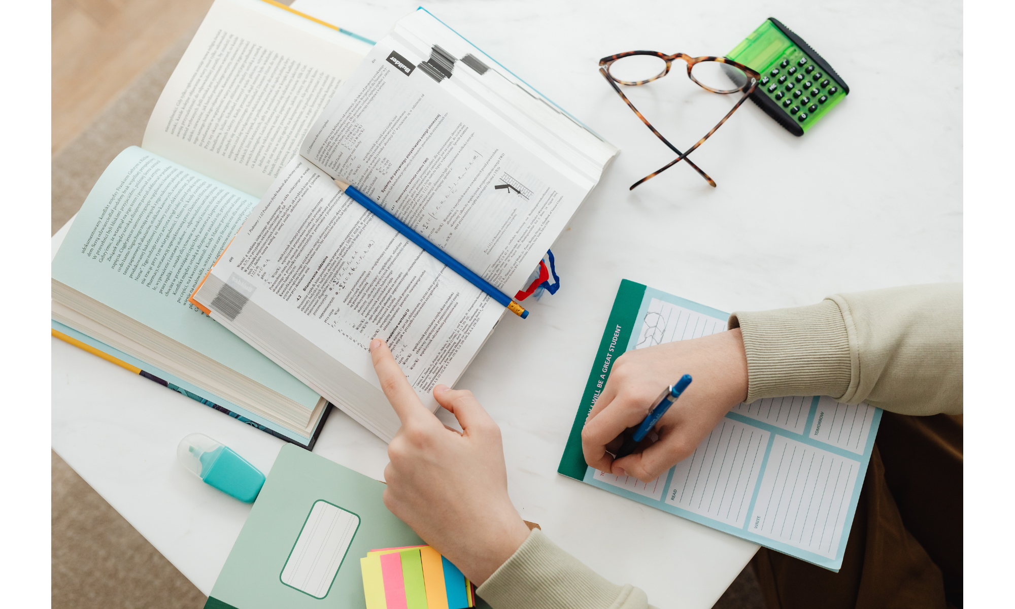 Person studying at a white table with open books, a blue pen, a green planner, a sheet of paper with survey questions, a small bottle of hand sanitizer, colorful sticky notes, eyeglasses, and a green calculator.