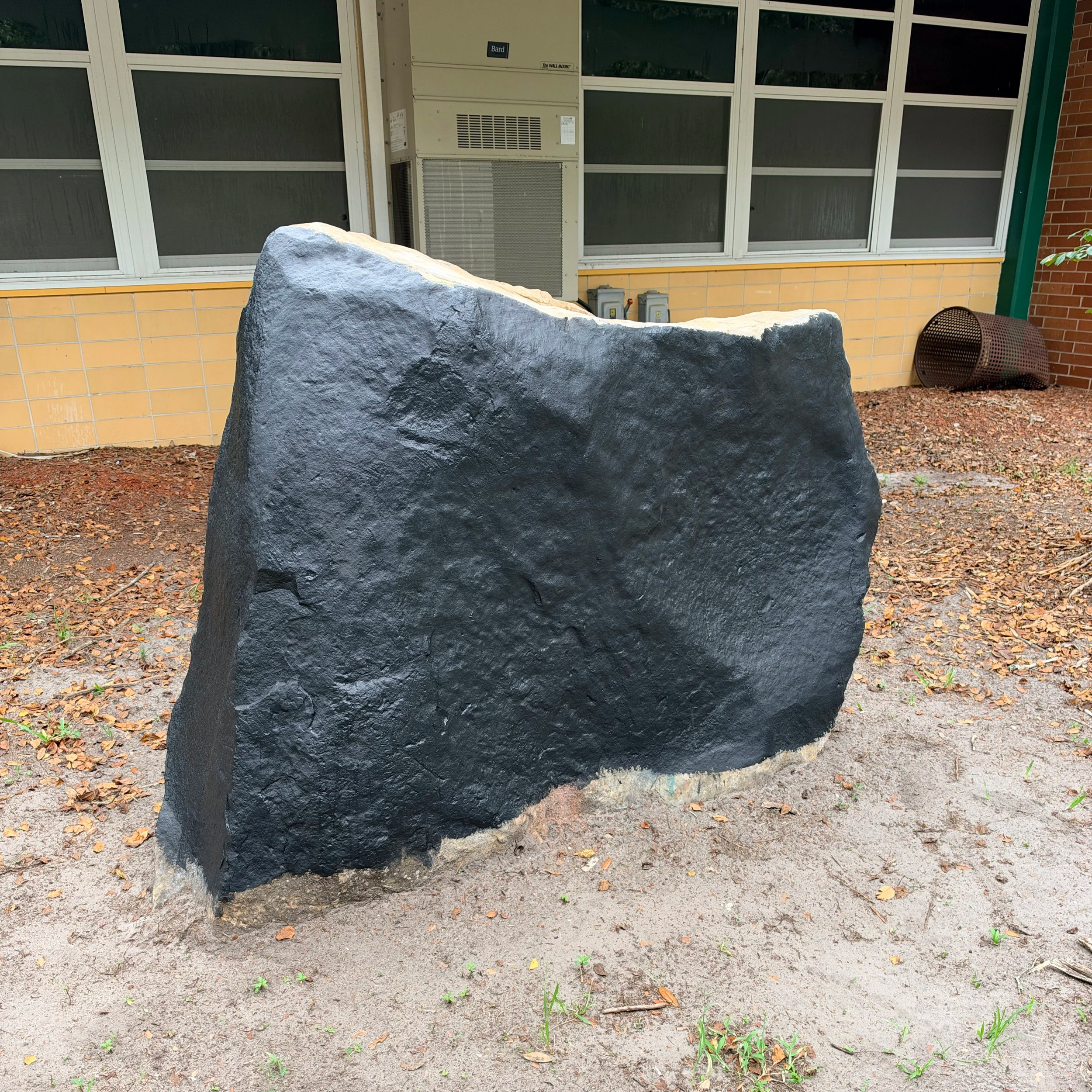 A large gray and black rock in front of a building with yellow tiles, large windows, and an air conditioning unit. There is some dirt and small green plants on the ground.