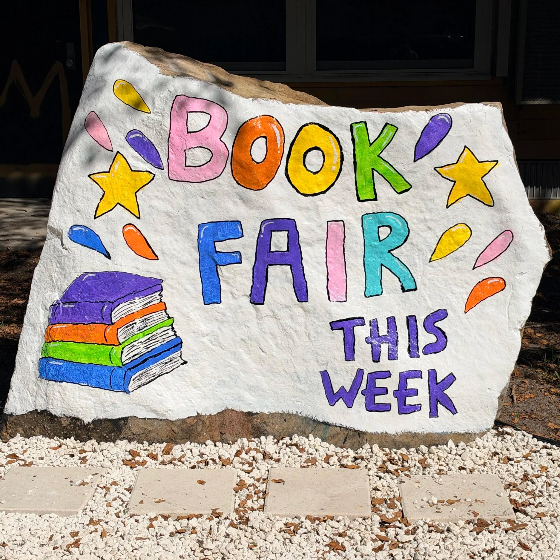 Colorful painted sign on a large rock with the words 'Book Fair' and illustrations of stars, water drops, and stacked books outside at night.