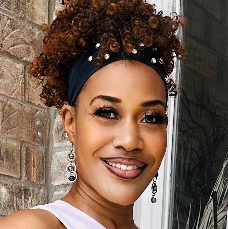 Close-up of a woman with curly hair, wearing earrings, and a black headband with white embellishments, smiling outdoors.
