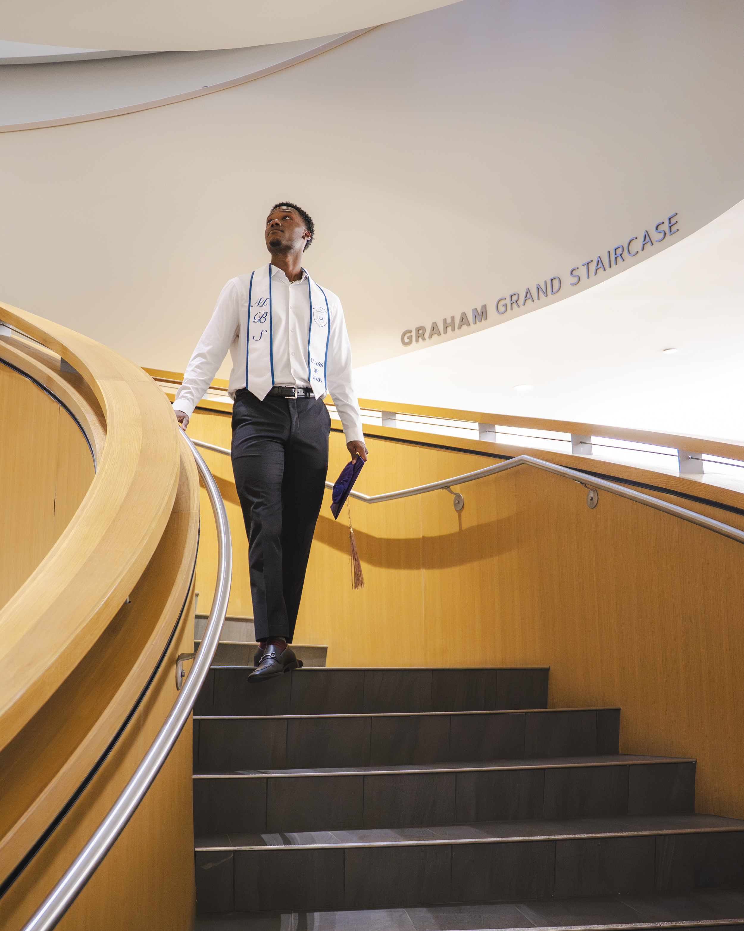 A man wearing a white shirt, dark pants, and black shoes walking down a staircase with a wooden handrail, holding a graduation cap with a maroon tassel in his right hand, in a modern building. The wall behind him is white with the sign 'GRAHAM GRAND 
