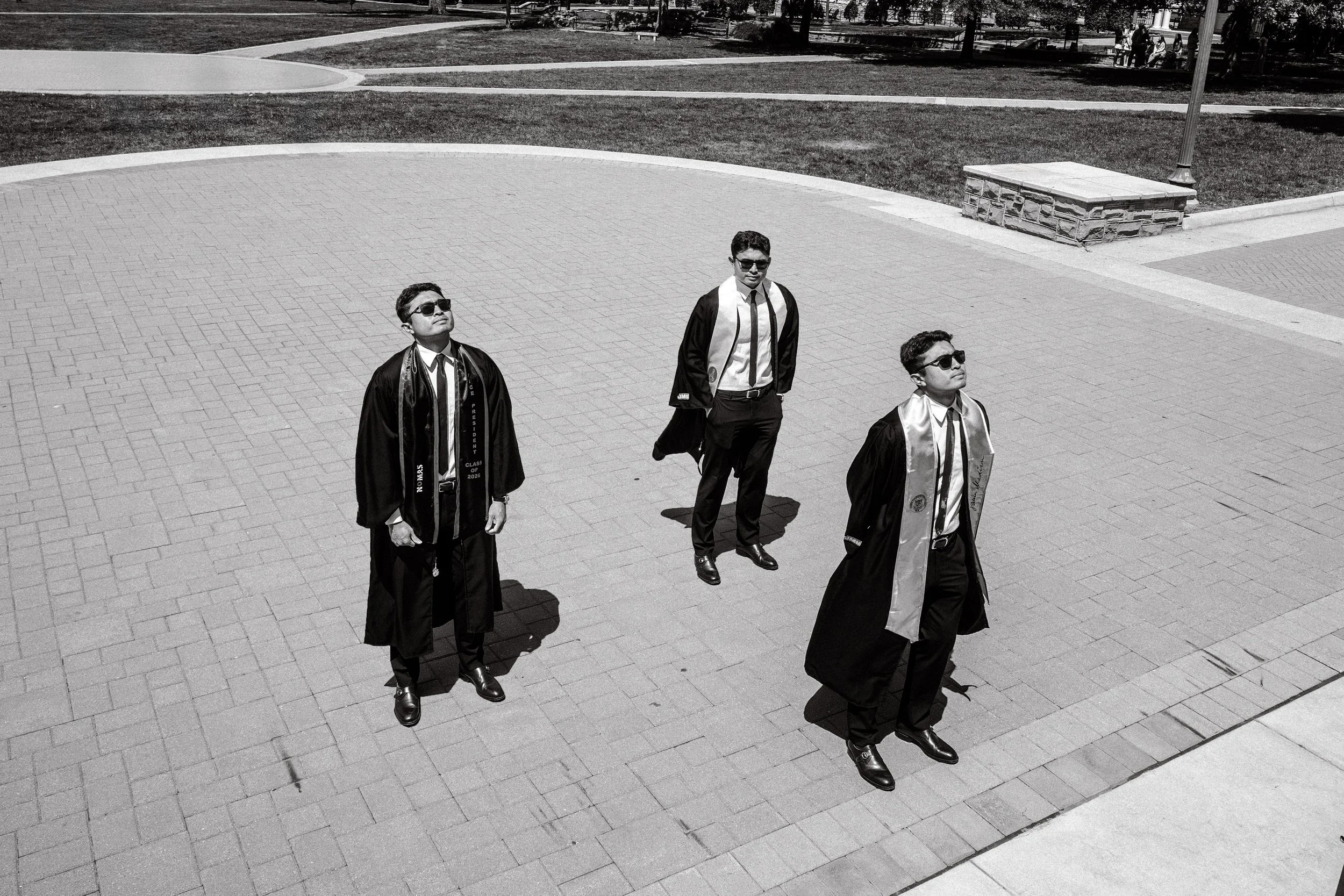 Three young men in graduation gowns and caps walking outdoors on a paved area, with trees and people in the background.
