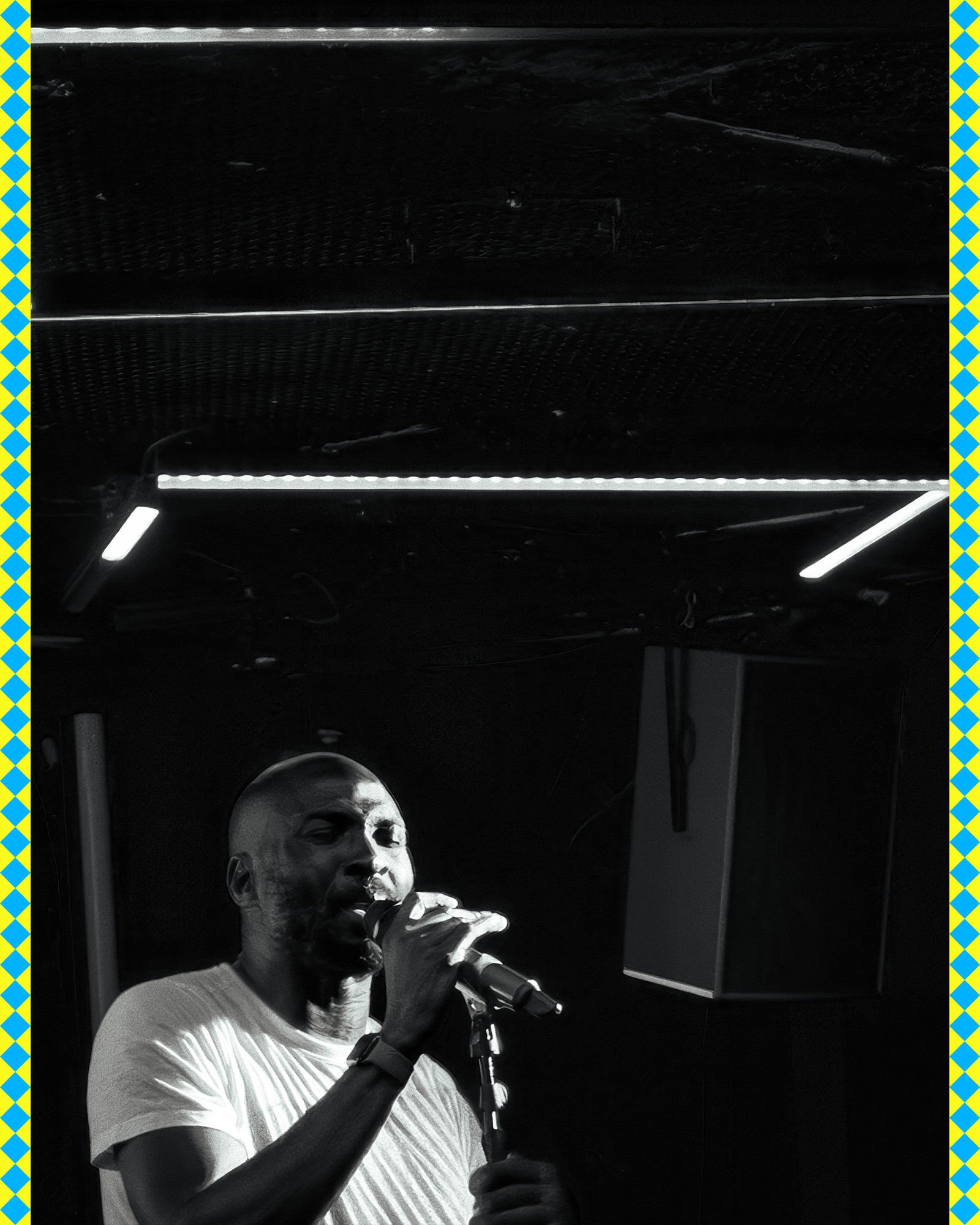 Black and white photo of a man singing into a microphone, wearing a striped t-shirt, with a dark background and spotlights overhead.