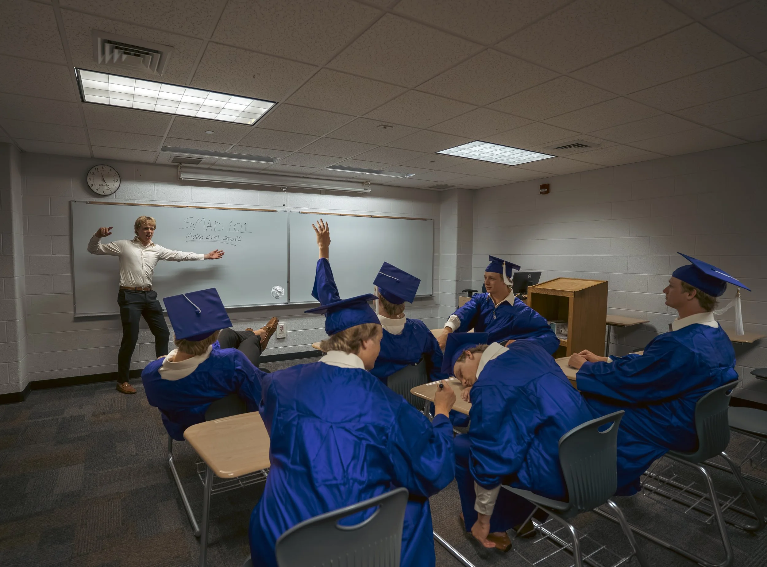 A classroom scene featuring a teacher engaging with students, some of whom are raising their hands, while others are seated at desks. The students are wearing blue graduation caps and gowns. The classroom has a whiteboard with writing that reads "SMA