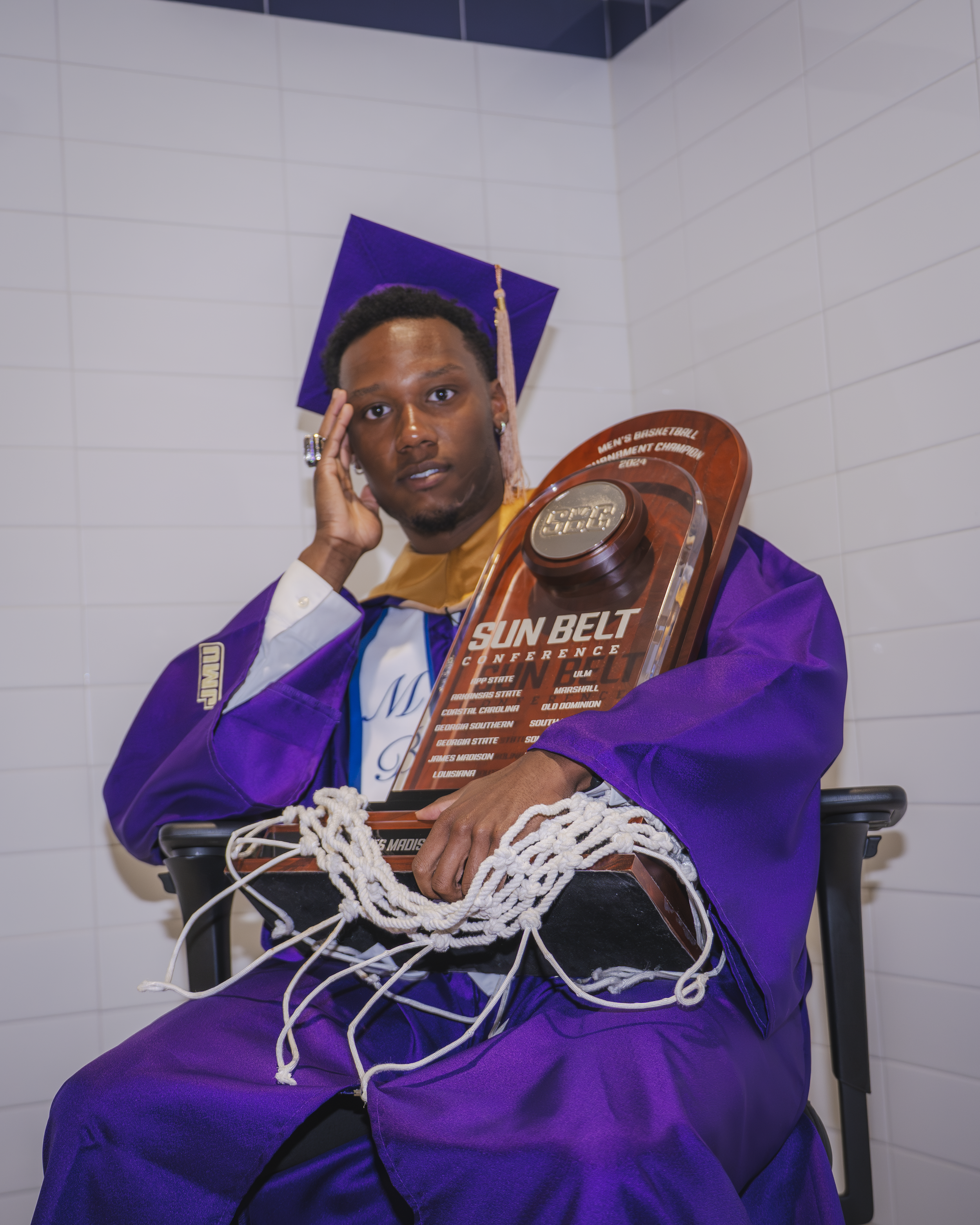 A young man wearing a purple graduation gown and cap holding a trophy with the text "Sun Belt Conference" and surrounded by a pile of white cords. He is sitting in a wheelchair against a white tiled wall, with a thoughtful expression and touching his