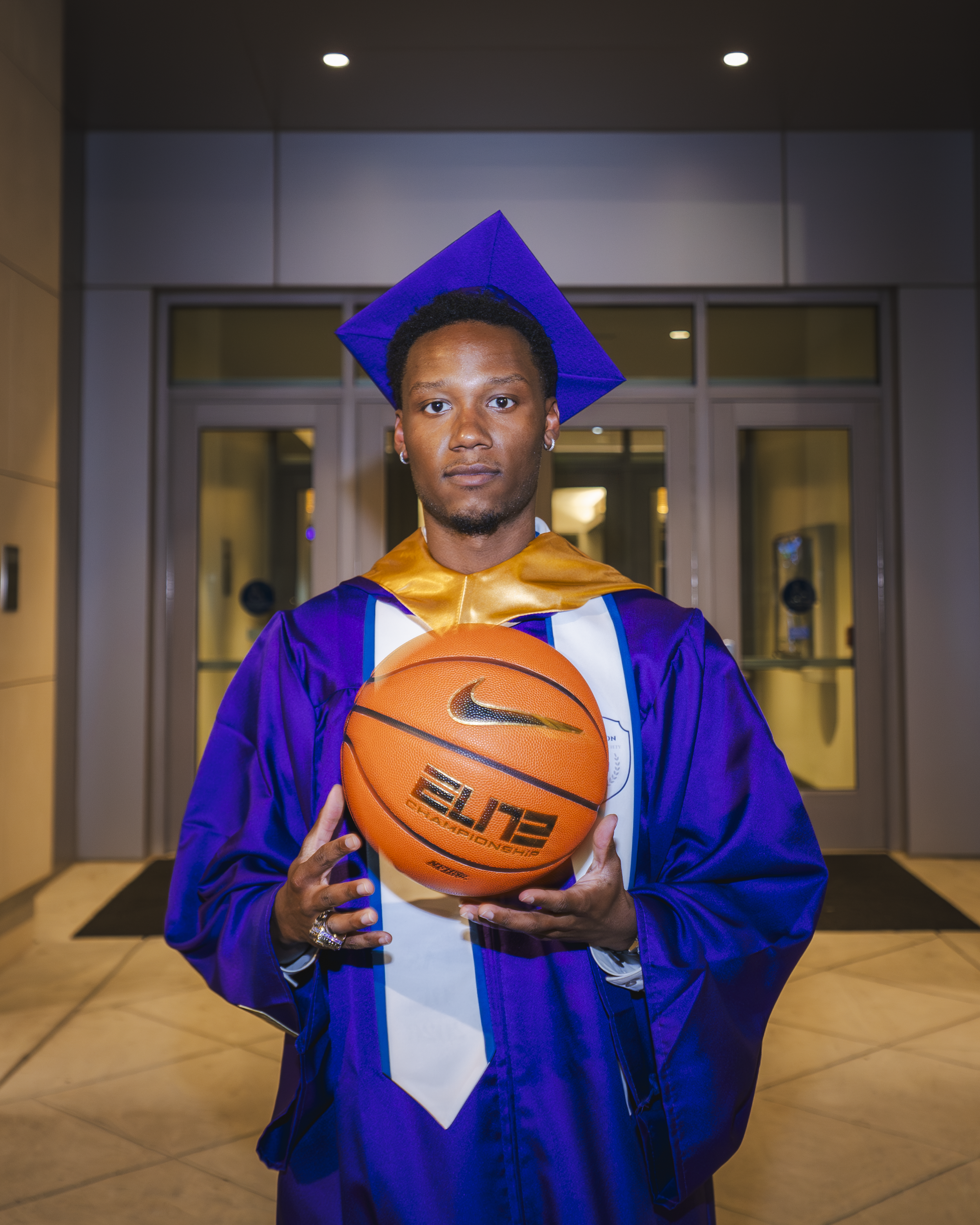 A young man wearing a blue graduation cap and gown holding an orange basketball with a Nike logo, standing in a modern indoor hallway.
