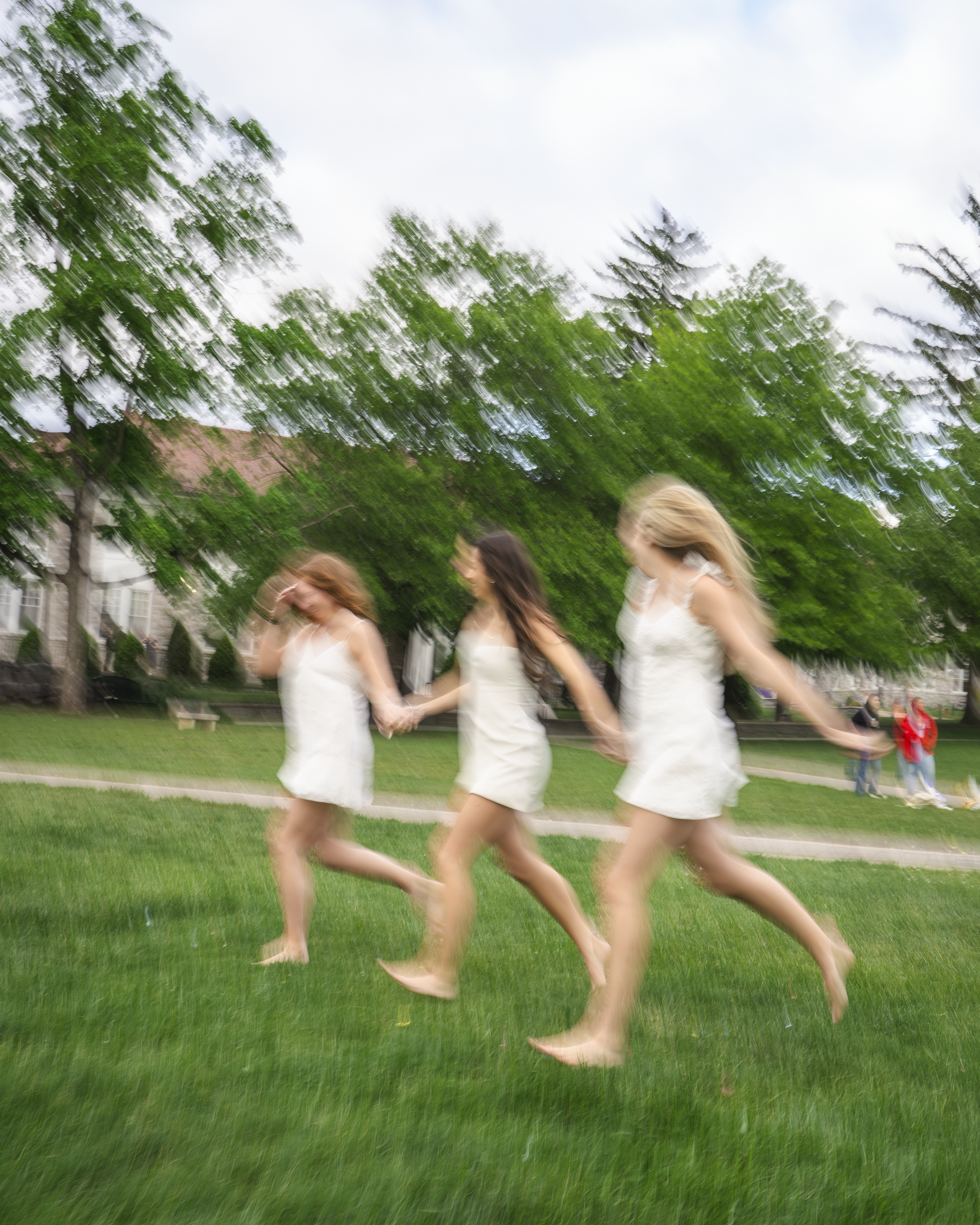 Four women in white dresses holding hands and running on a grassy lawn, with large green trees and a building in the background, under a cloudy sky.