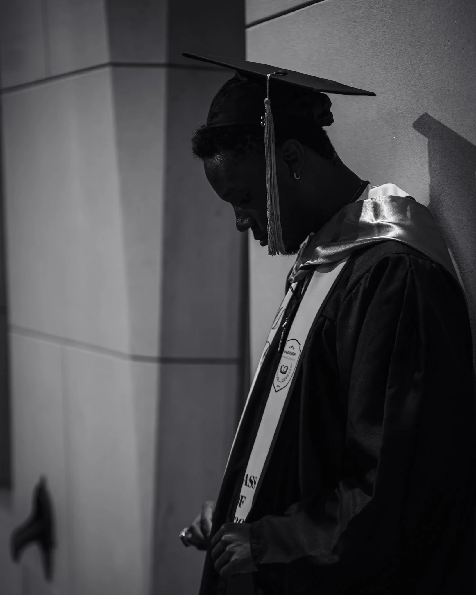 A person at a graduation ceremony wearing a cap and gown, standing against a wall, holding a diploma. The image is in black and white.
