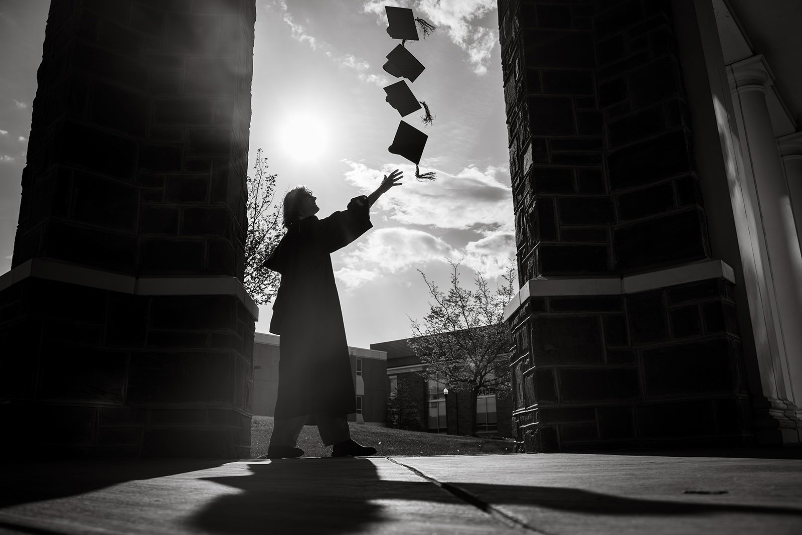 A woman in a graduation gown is tossing her cap into the air outdoors with a bright sun behind her, framed by dark brick columns.
