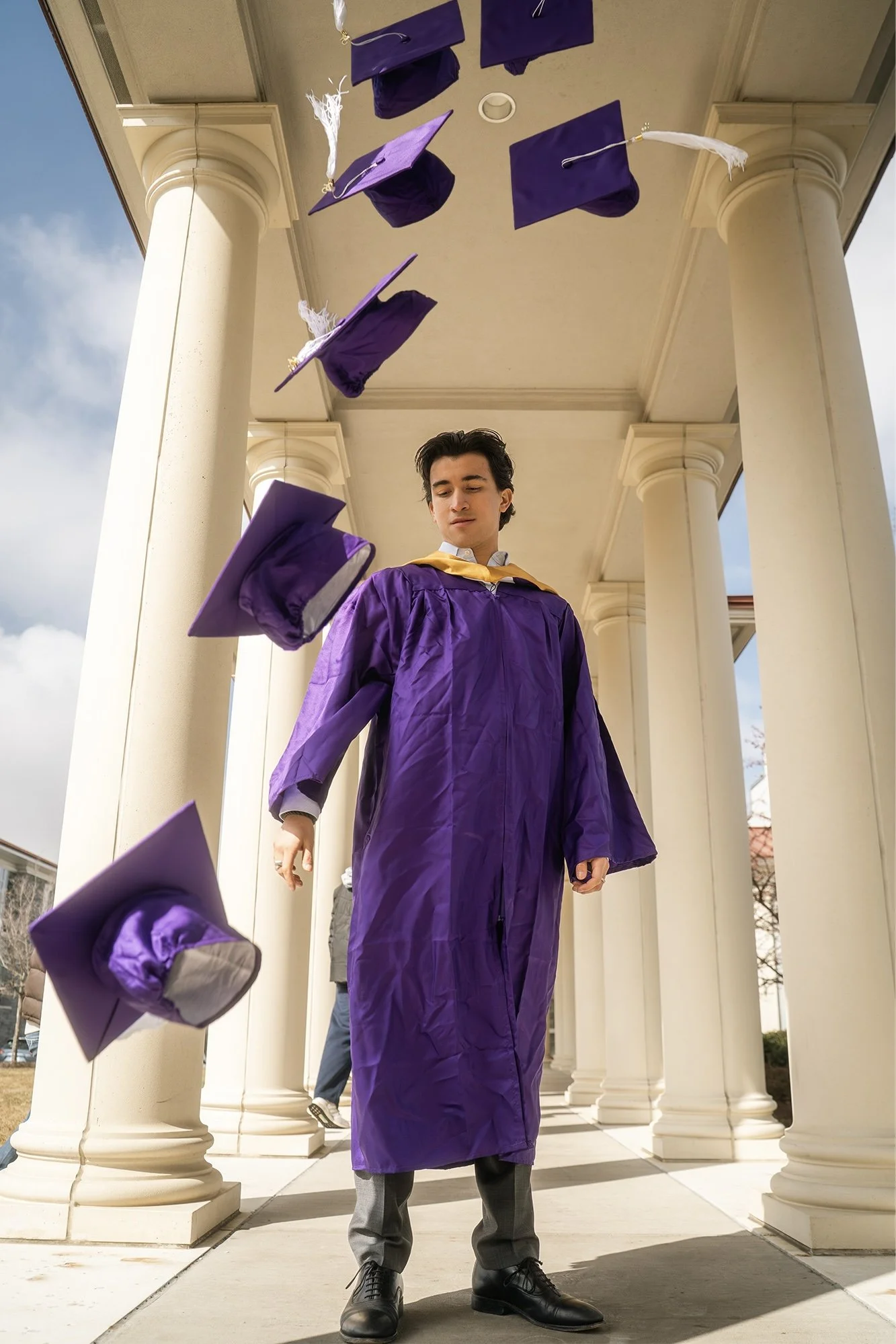 A young man in a purple graduation gown and cap stands outdoors under a colonnade with falling mortarboards around him, facing forward with a neutral expression.
