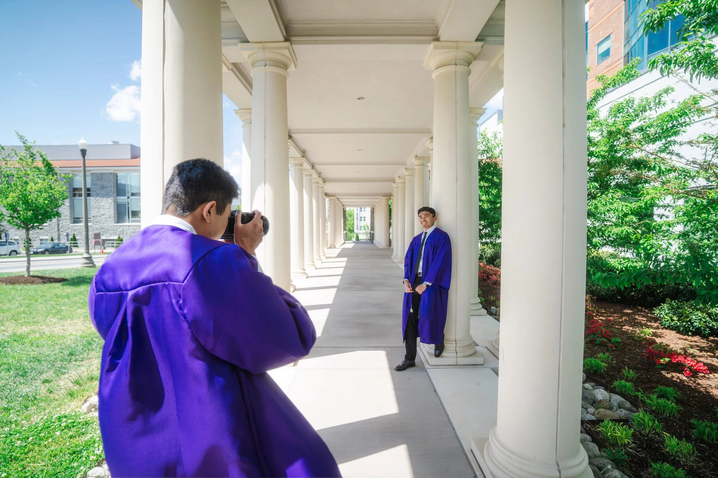 A young man in a graduation gown taking a photo of another young man in a blue graduation gown, who is posing against white columns outside a building with trees and greenery.