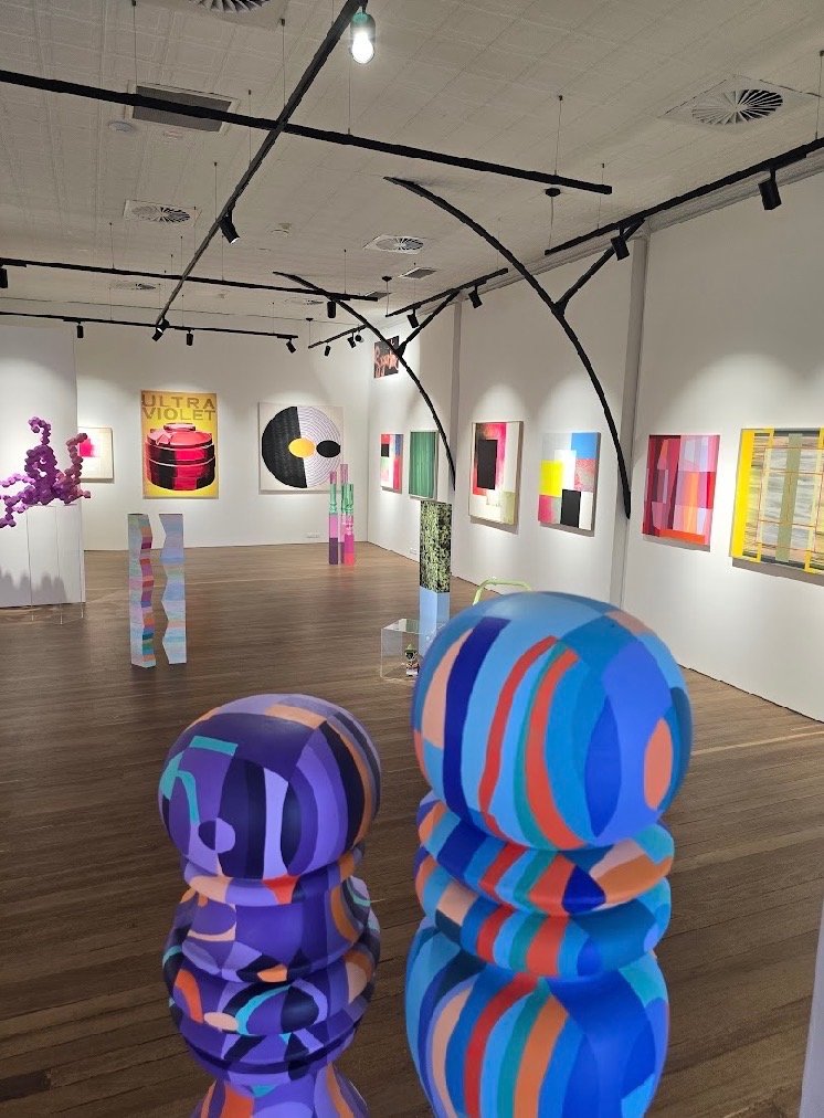 An art gallery with colorful abstract paintings on white walls and sculptures on the wooden floor, including striped, spherical, and organic shapes.