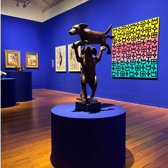 Sculpture of a child holding a dog overhead in an art gallery with colorful geometric paintings on blue walls.