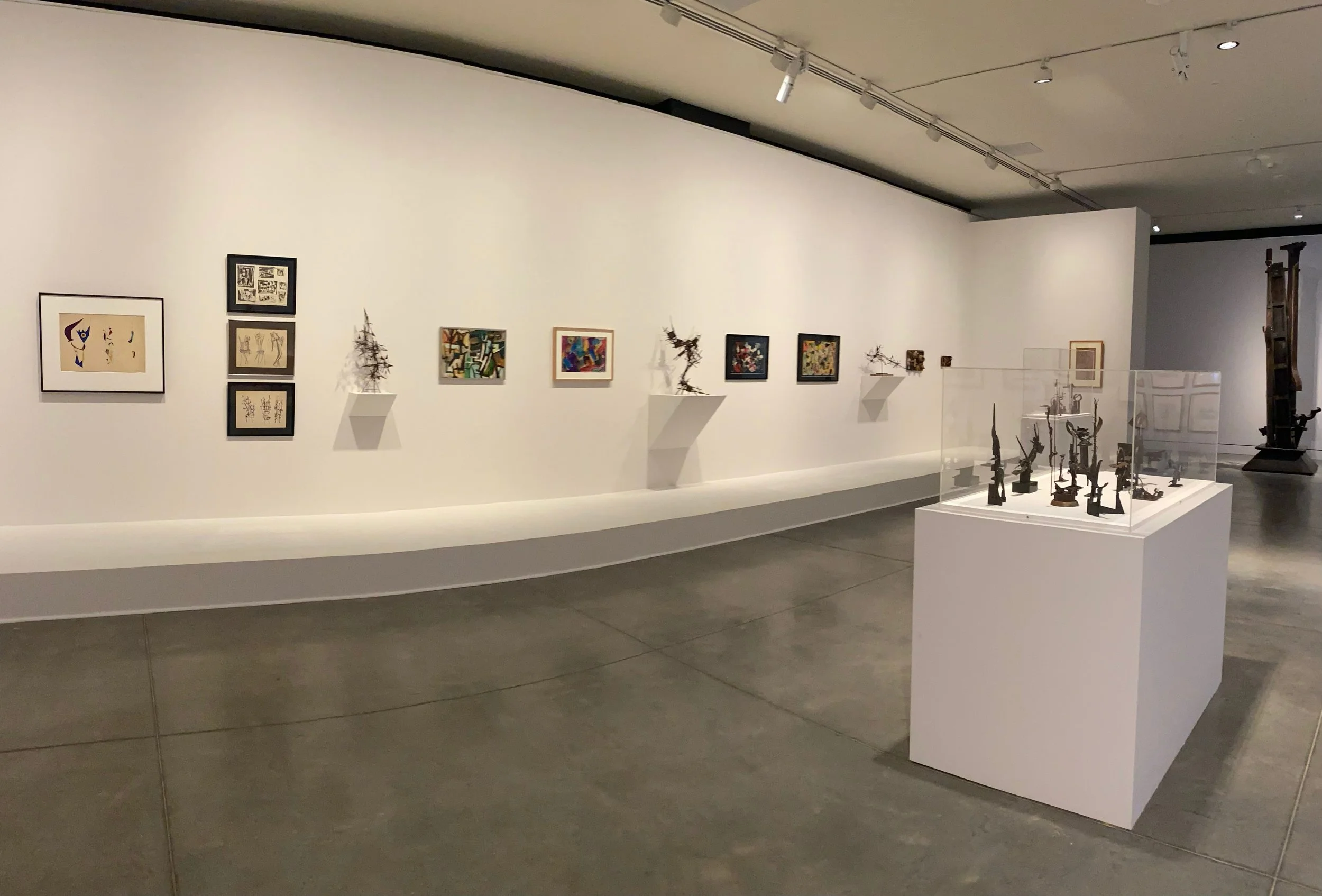 Art gallery with various framed artwork on a white wall, small sculptures on wall-mounted shelves, and art pieces displayed in a glass case, with a large sculpture in the background.