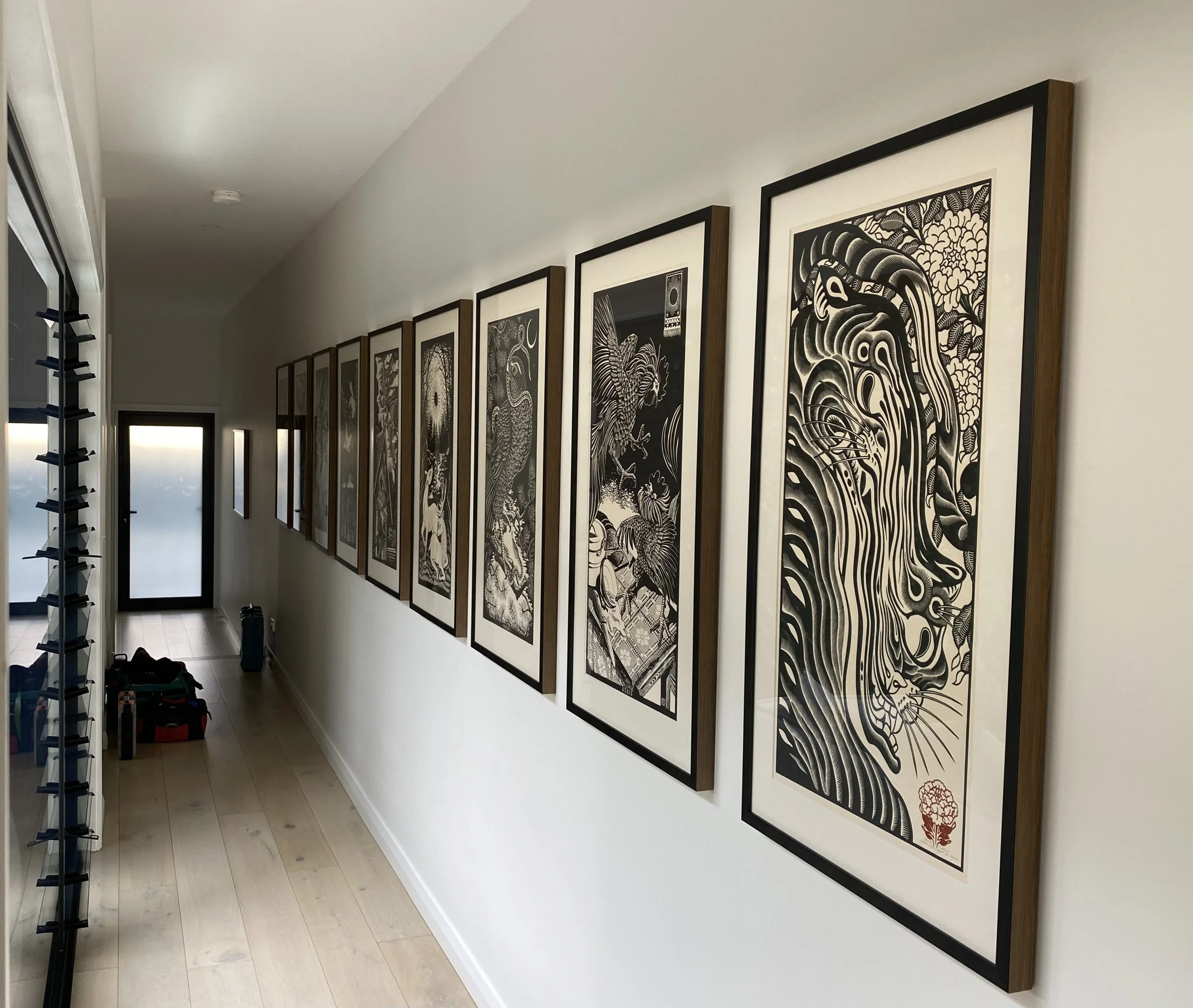 A hallway with a series of framed black-and-white Asian-style artwork depicting various animals and nature scenes, along with a row of shoes and bags on the floor near a frosted glass door.
