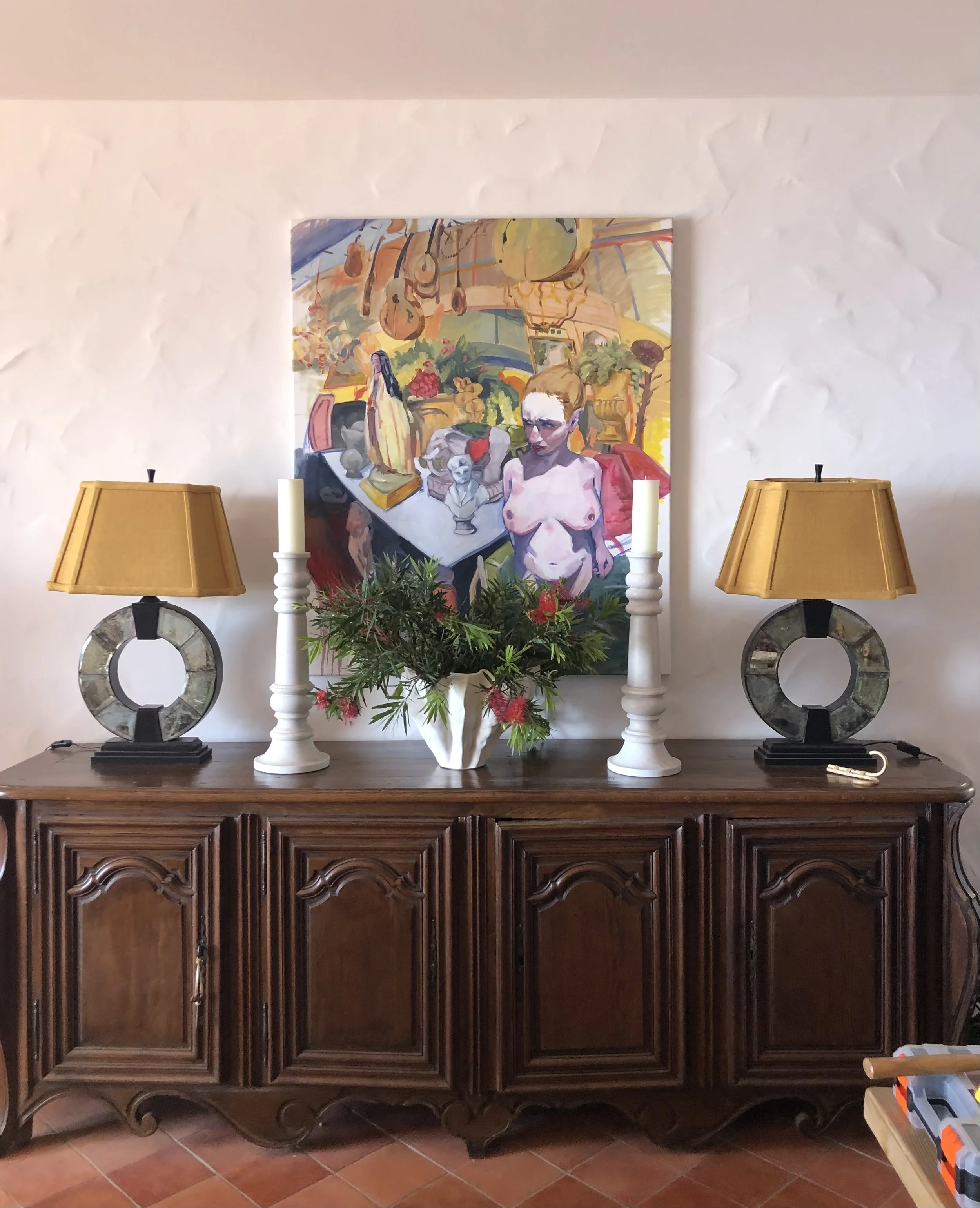 A wooden sideboard with two yellow lamps, candles, greenery, and a colorful painting behind.