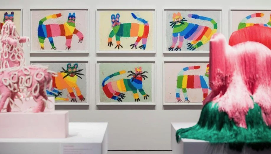 Gallery wall with colorful, abstract animal paintings, including dogs, cats, and other creatures, with two pink and green sculptural art pieces in the foreground.