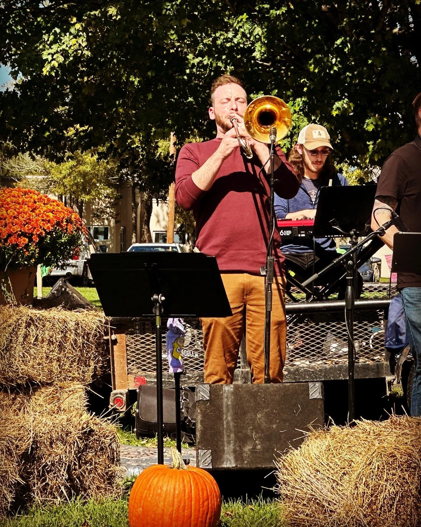A man with a beard, wearing a red long-sleeve shirt and brown pants, is playing a trumpet outdoors during the daytime. Behind him, another musician is standing at a keyboard. There are hay bales, pumpkins, and trees in the background.