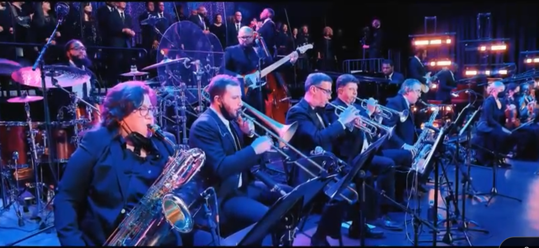 Jazz band performing live on stage with musicians playing brass, woodwind, and percussion instruments.