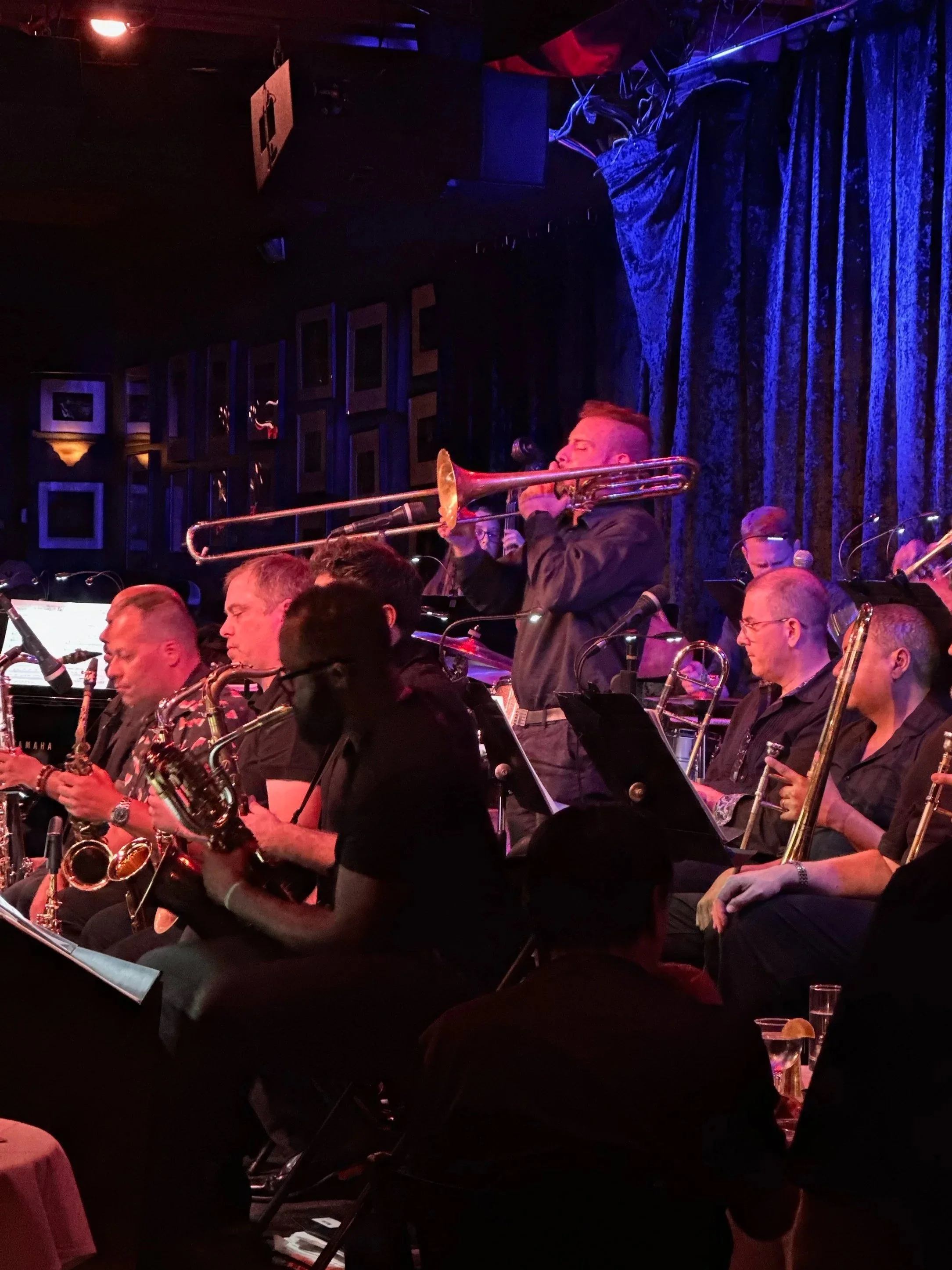 A jazz band performing on stage with a trombone player standing and playing, while other musicians sit and play saxophones and other instruments. The stage has dark curtains and framed pictures on the wall behind them.