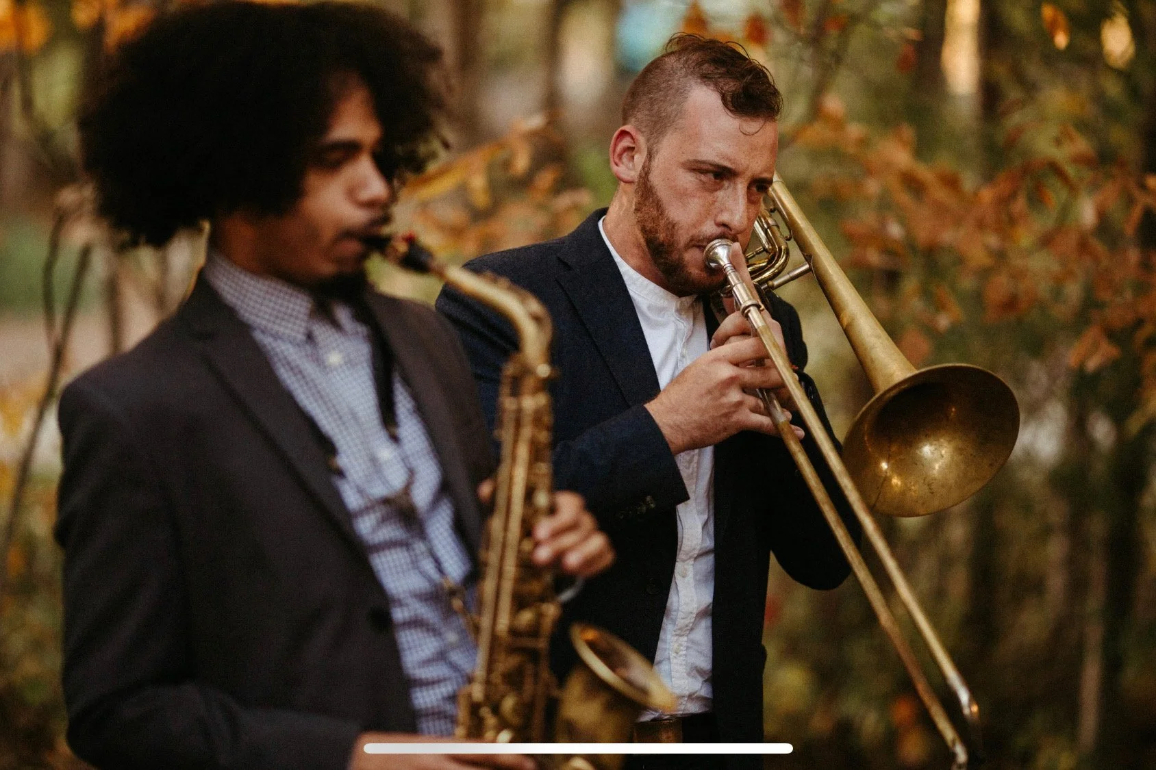 Two men playing saxophone and trombone outdoors on a fall day, surrounded by trees with autumn leaves.