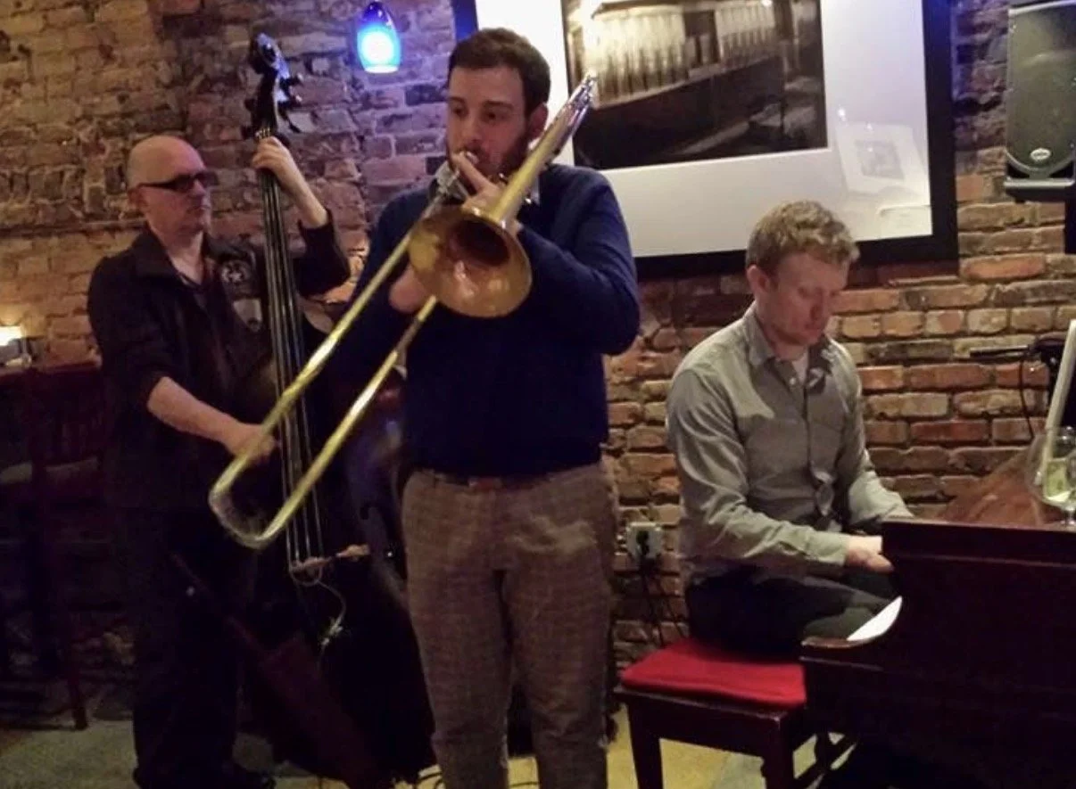 Jazz musicians performing in a cozy venue with brick walls, including a trombone player, a double bassist, and a pianist.