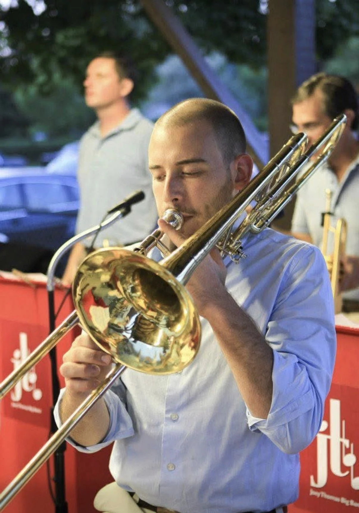 A man playing a trombone at an outdoor concert, with other musicians in the background.