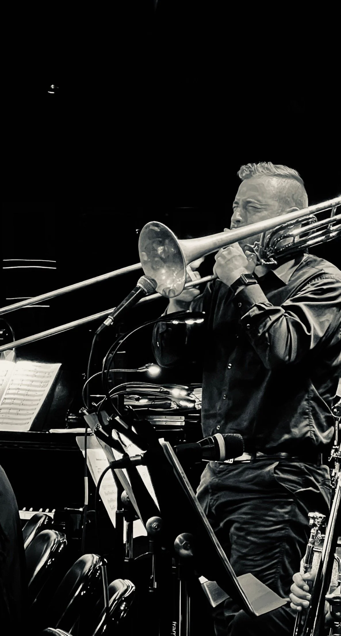 A musician playing a trombone during a live performance, with sheet music and other instruments visible in the background, in black and white.