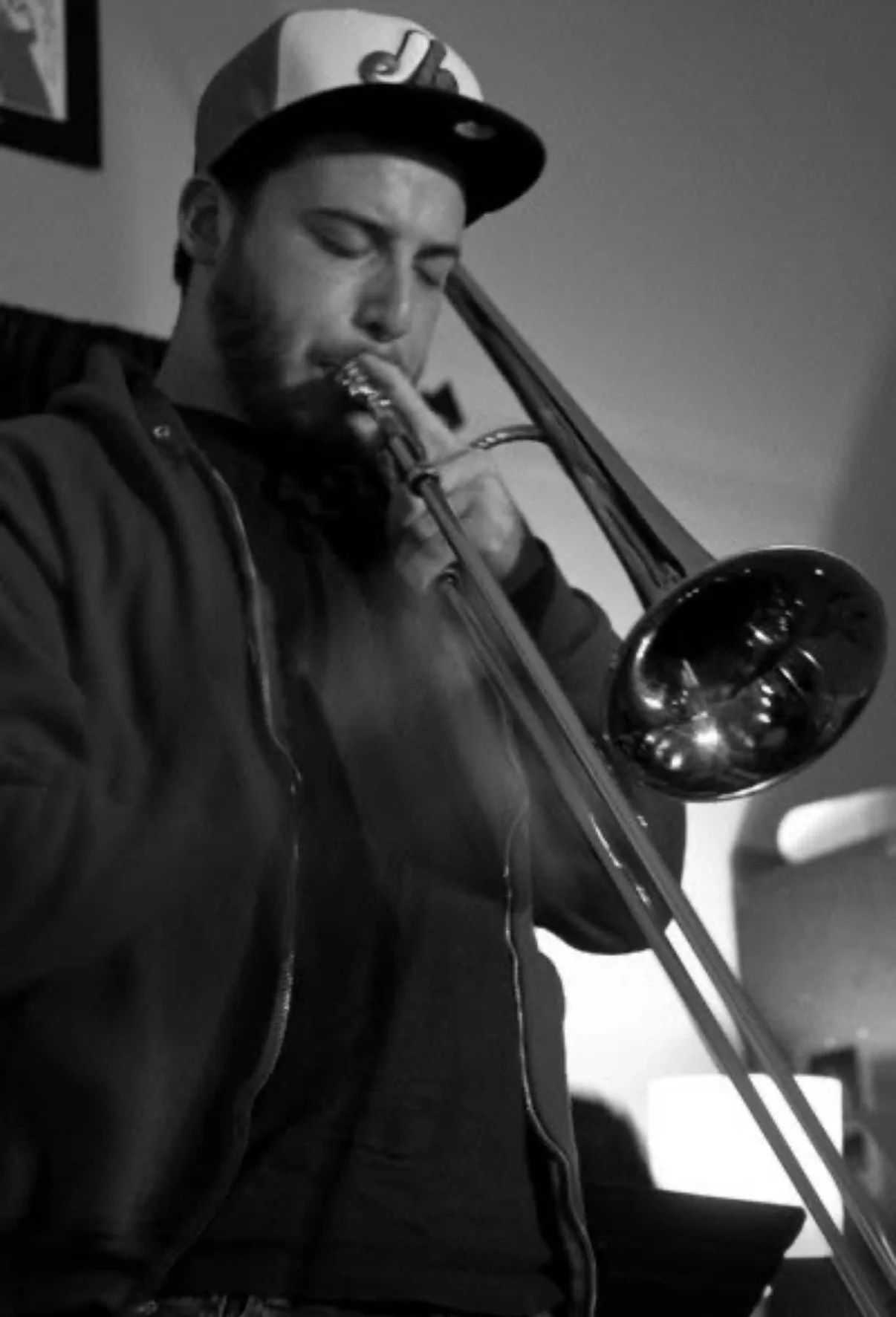 A young man wearing a baseball cap is playing a trombone, dressed in a dark jacket, in a room with blurred background.
