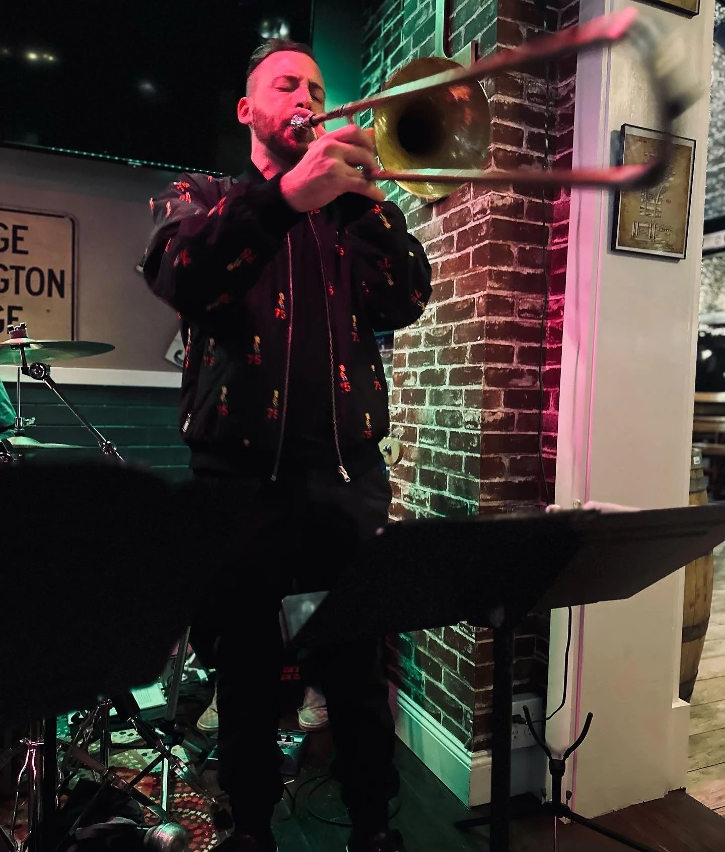 Man playing trombone at a live music venue with a brick wall background and music stand in front.