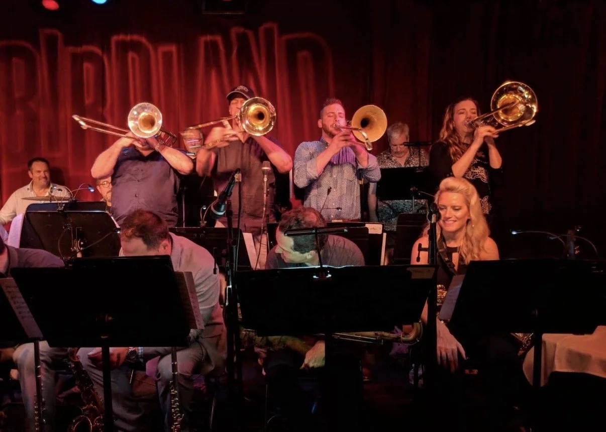 Musicians playing brass and wind instruments on stage during a jazz concert at a red velvet curtain venue.