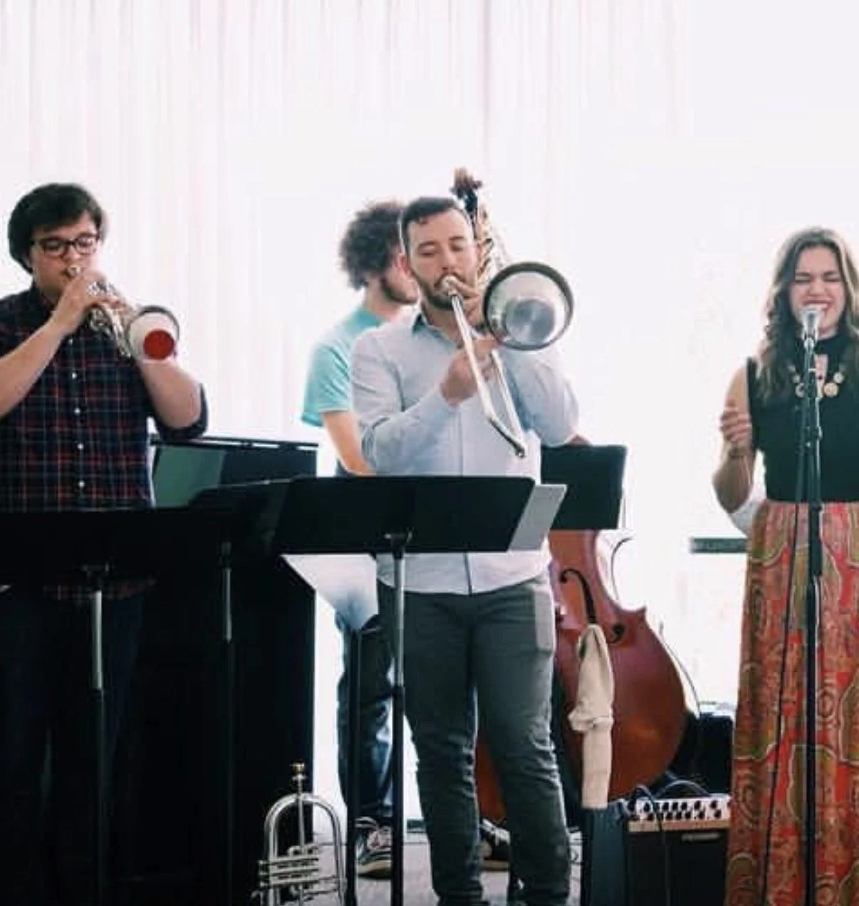 A group of five musicians playing different instruments including a trumpet, trombone, and vocals, in a bright room with a large window.