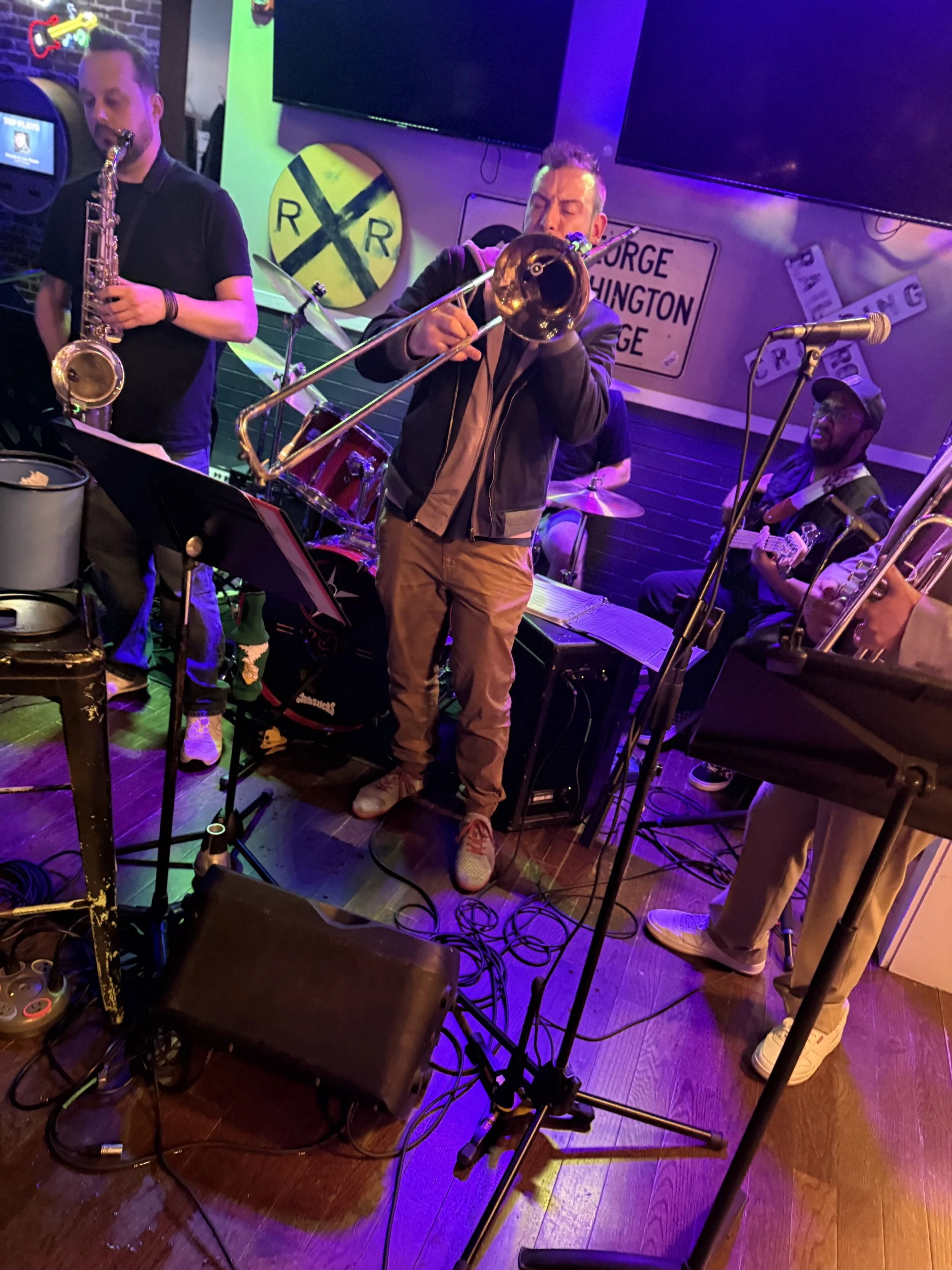 Band performing live on stage at a bar or club, with musicians playing guitar, saxophone, trombone, and drums.