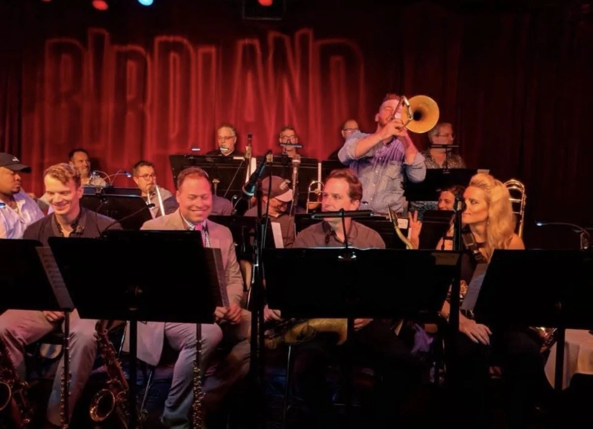 Jazz band performing on stage with a prominent red curtain background and the words 'BROADLAND' in neon lights, featuring musicians playing saxophone, trumpet, and other instruments.