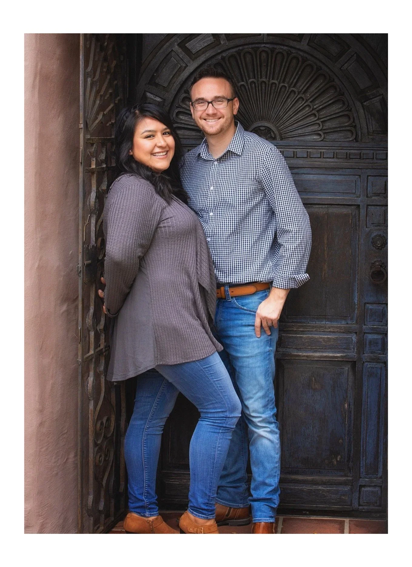 A smiling couple standing close to each other in front of a dark ornate wooden door with an arch design. The woman has long black hair, is wearing a gray ribbed sweater, and jeans, and is standing slightly in front of the man. The man has short dark hair, glasses, a checkered shirt, and blue jeans, and has his arm around the woman's waist.